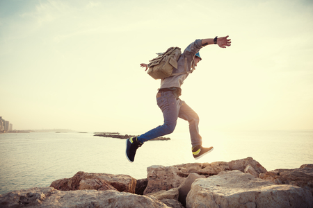 Brave man with backpack running over rocks near ocean in sunsetの写真素材