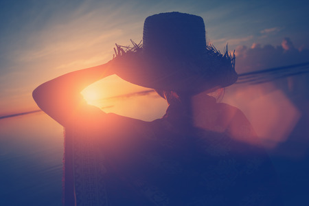 Womans silhouette in hat looking at sunrise at the ocean, lens flare effect and vintage colorの写真素材