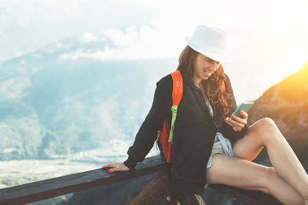Smiling young woman with backpack and white hat sitting in mountains and typing message on her mobile phone, outdoorsの写真素材