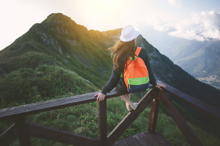 Young traveling woman wearing hat and backpack sitting high on the top of the mountain with waving hair, evening with sunset, outdoorsの写真素材