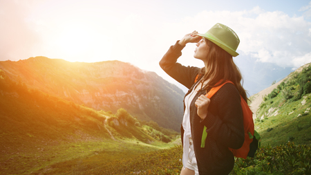 Woman traveling along mountain meadows. With small backpack and sun hat. Evening with sunset, outdoorsの写真素材