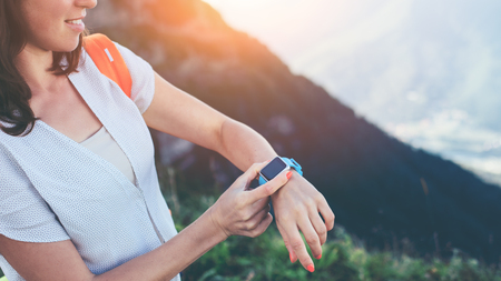 Young smiling woman adjusting smart watches on her wrist, while trekking and walking along mountain meadows, outdoorsの写真素材