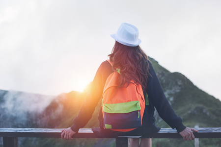 Traveling woman wearing hat and backpack sitting high on the top of the mountain with waving hair, evening with sunset, outdoorsの写真素材