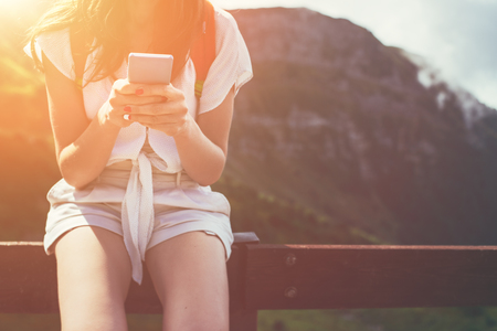 Young woman sitting on the fence with mobile phone. High mountains touristic path. Digital communication. Texting with friends in social networkの写真素材