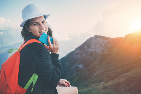 Young woman wearing a hat and backpack using the phone, sitting high in beautiful mountain reserve...
