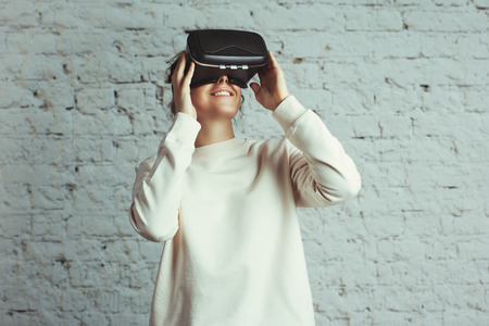 Handsome young woman wearing virtual headset. Smiling hipster using VR glasses. Blank sweater. White bricks wall background, studio indoorsの写真素材