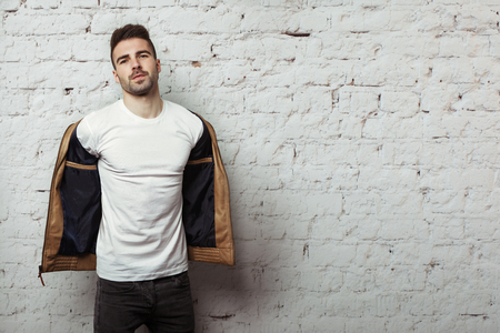 Handsome man in blank t-shirt taking off his leather jacket, white bricks wall background, studio indoorsの写真素材