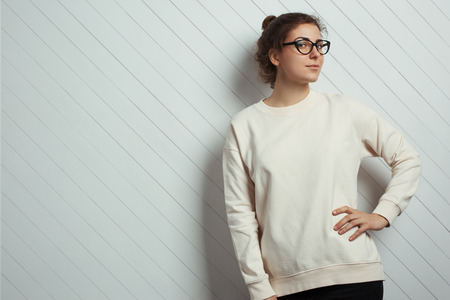 Portrait of charming woman hipster wearing blank sweater and eye glasses. White wooden wall with background, studio indoorsの写真素材