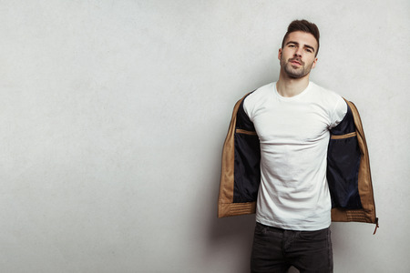 Handsome man in blank t-shirt taking off his leather jacket, grey concrete wall background, studio indoorsの写真素材