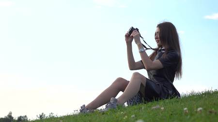 Beautiful brunette girl in black dress sitting on the lawn and making photos with her camera....