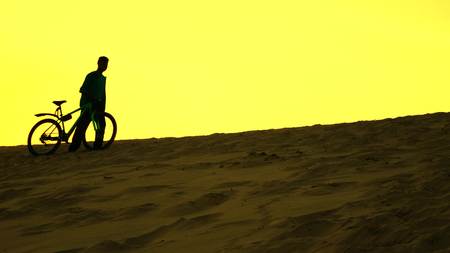 Silhouette of lonely biker walking with his bike along sand dune, yellow colors