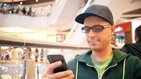 Young caucasian man wearing cap and black rim glasses taps on his mobile phone touchscreenの写真素材