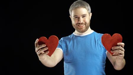 Smiling bearded man in blue tshirt holds two red heart shapes. Love, romance, dating, relationship...