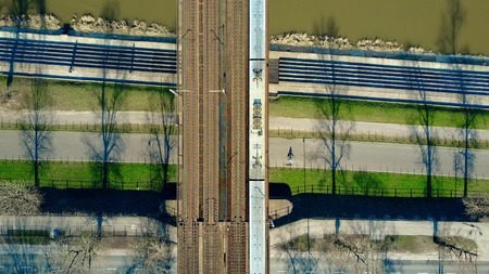 Aerial shot of passenger train moving on railway bridge across the river and bicycle road, top view....