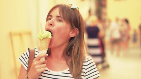 Young beautiful woman eating ice cream and walking along crowded tourist street, summer warm colors