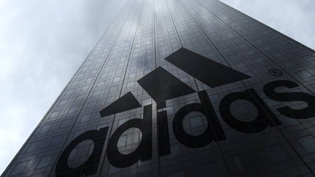 Adidas inscription and logo on a skyscraper facade reflecting clouds. Editorial 3D rendering