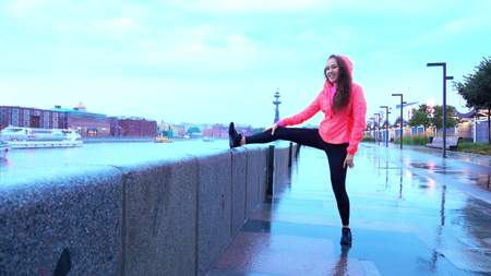 Young mixed race athletic woman warming up before running along city park embankment