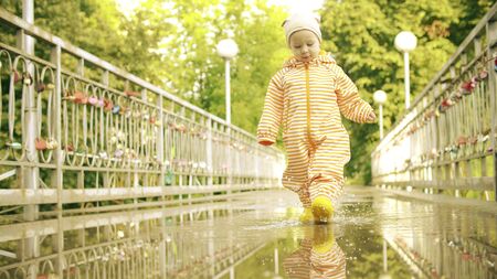 Funny little girl in orange waterproof raincoat and rubber rain boots walks on puddle after rain