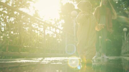 Happy baby girl in waterproof raincoat and rain boots walks on puddles, slow motion shotの写真素材