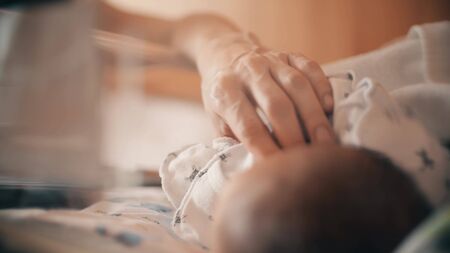 Mom touches her newborn baby in a transparent hospital bassinetの写真素材