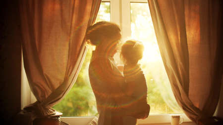 Mom holding and hugging her little daughter near the window at home, family timeの写真素材