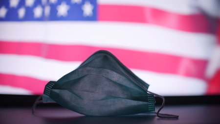 Disposable mask against blurred flag of the USA. American COVID-19 outbreak conceptの写真素材