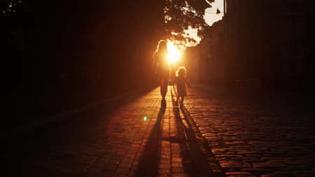 Silhouette of unknown mom and little daughter walking along the street paved with cobblestone in the eveningの写真素材