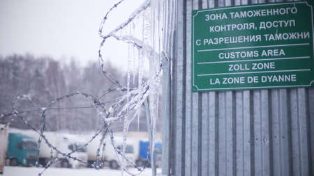 Blurred trucks behind barbed wire fence and customs area sign in Russia, same text in different langugesの写真素材