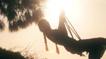 Silhouette of a slim beautiful woman in dress swinging on the swing against shining sunの写真素材