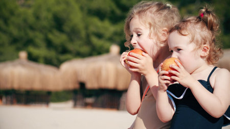 Two little girls eat apples on the beach together. Healthy eating conceptの写真素材