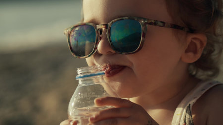Little baby girl wearing sunglasses drinks water from plastic bottle on the beachの写真素材