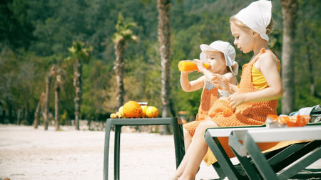 Adorable little sisters wearing orange clothes drink fresh orange juice on the beachの写真素材
