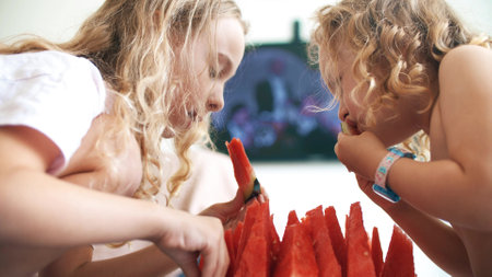 Little sisters eat juicy watermelon slices and watch TV at homeの写真素材