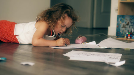 Little curly girl draws a picture with a black felt pen on the floor at homeの写真素材