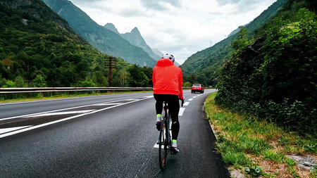 Follow camera view of a cyclist in red jacket riding on the road to Oz Lake in the French Alps, surrounded by dramatic green mountains and cloudy sky, symbolizing endurance and freedomの写真素材