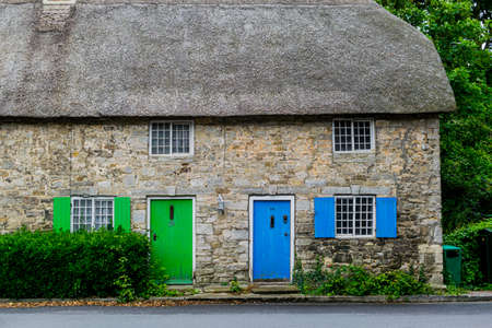 West Lulworth, United Kingdom - 19 July 2020: Beautiful thatched cottage house with blue and green coloured door and shutters, unique Dorset colorfull house decorations by Main Roadのeditorial素材