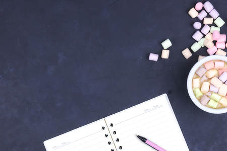 Cup with coffee and marshmallows, blank open notebook with pen on black concrete background. View...