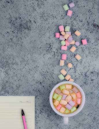 Cup with coffee and marshmallows, blank open notebook with pen on abstract blue background. Top...