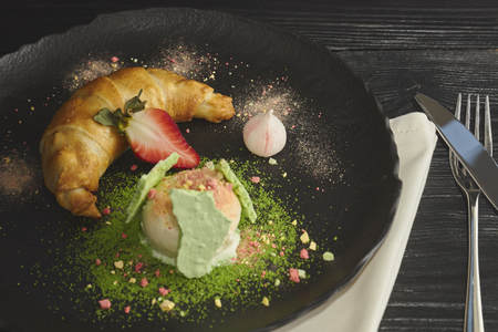 Slice of apple strudel with a scoop of vanilla ice cream on a black plate, on a wooden black table, decorated with meringue and strawberries. close-up. horizontal view of bakedの写真素材