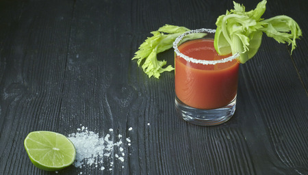Cocktail Bloody Mary with lime and celery, salt and snacks in a glass on a black wooden table.の写真素材