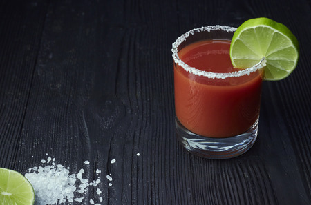Cocktail Bloody Mary with lime and celery, salt and snacks in a glass on a black wooden table.の写真素材