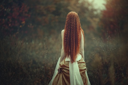 A beautiful young woman with very long red hair as a witch walks through the autumn forest. Back view. Creative colors and Artistic processing.の写真素材