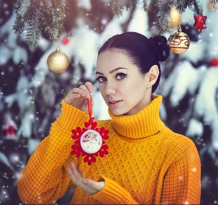 A beautiful girl with blue eyes in a yellow sweater is holding a New Years toy under a snow-covered tree. A girl in fairy-tale New Years decorations. Magic winter forest.の写真素材