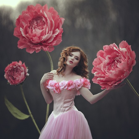 Beautiful sensual girl with red hair in a fairy pink dress posing near giant peony pink flowers. The...