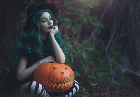 Halloween witch with green hair holding a carved scary pumpkin. Beautiful young girl in a black hat,...