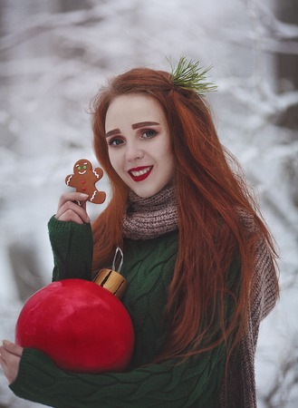 Red-haired long-haired smiling girl with a huge Christmas red ball eating ginger cookies. A young woman with red hair walks in a snowy forest.の写真素材