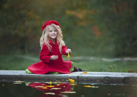 A little blonde girl in a red coat and beret throws leaves into the water. Girl sitting by the water. Autumn photography.の写真素材
