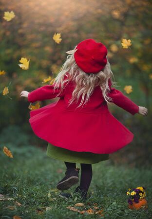 A little blond girl in a red coat and beret is spinning in yellow leaves in the autumn forest. Girl with pumpkin. Autumn photography.の写真素材