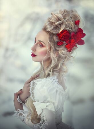 Portrait of an old-fashioned blonde woman with a medieval hairstyle of the XVI century. Her hair is decorated with red flowers. Powdered wig.の写真素材