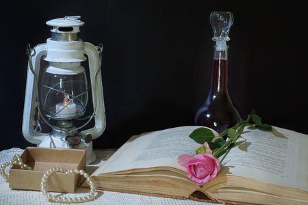 Horizontal still life on a black background with a book, an old lantern and a bottle of wine, a pink rose lies between the pages, next to a set of pearl jewelry in a cardboard box.の写真素材